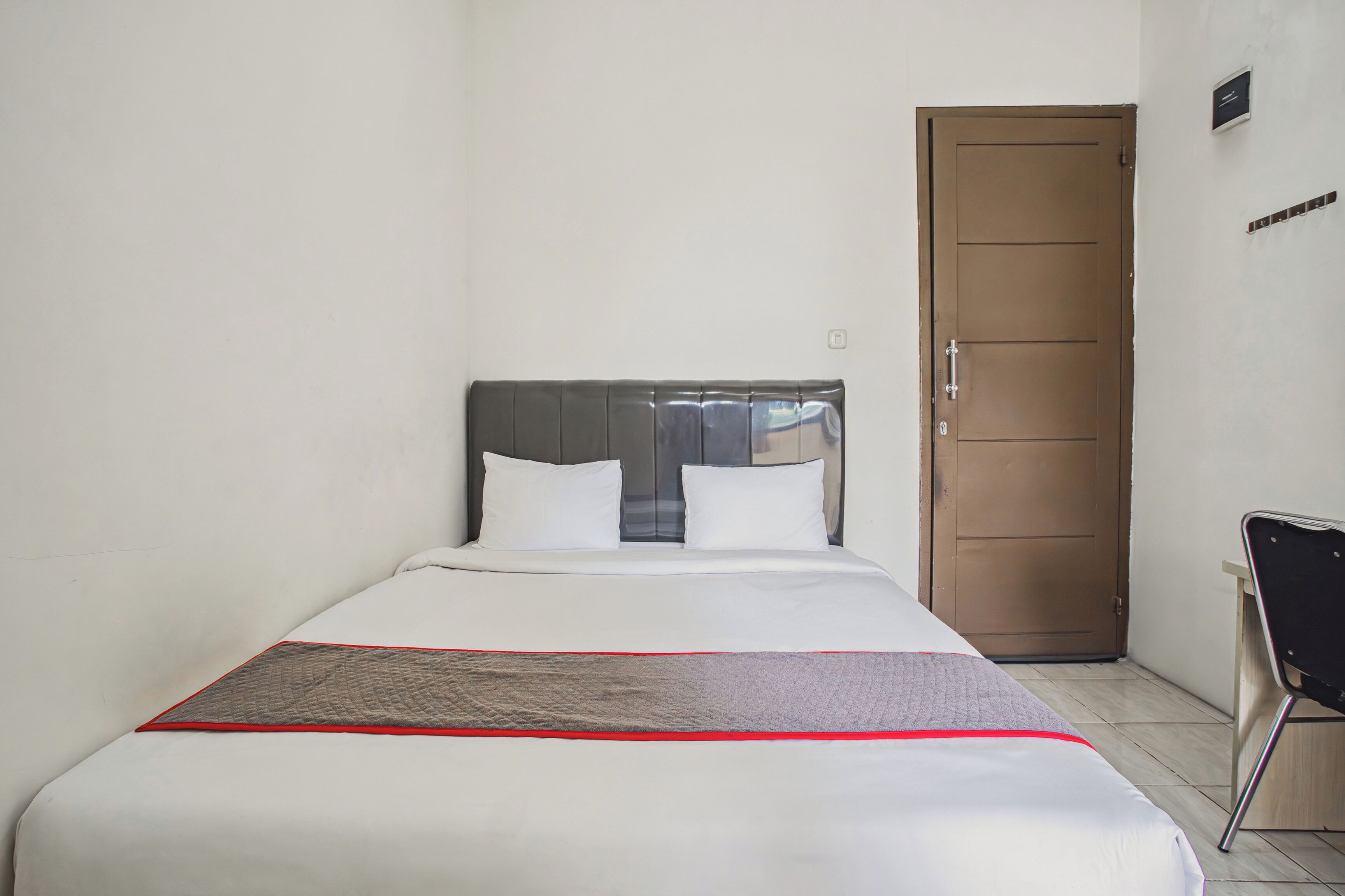 A neatly made bed with white bedding and a red accent, a closed wooden door, and a small desk with a chair.