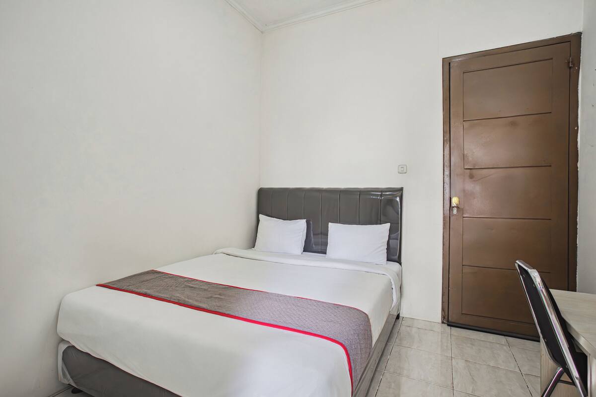 A neatly made bed with a grey headboard, a brown door, and a chair in a simple room with white walls.