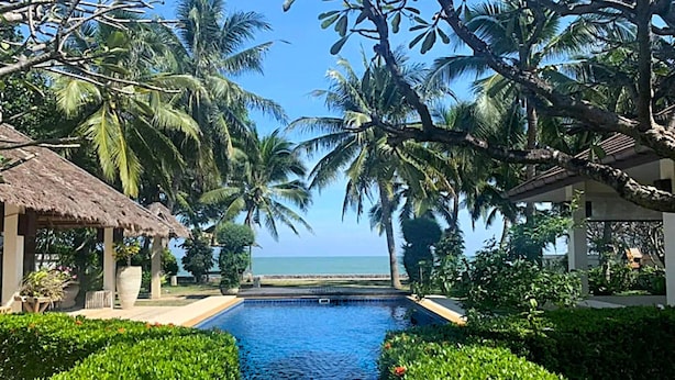 A pool with a view of the ocean, surrounded by palm trees and a thatched-roof structure.