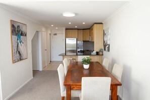 A dining area with a wooden table and chairs, a potted plant, and a kitchen in the background.