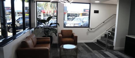 A modern waiting area with a sofa, armchair, and round table, large windows with a view of parked cars, and a staircase.