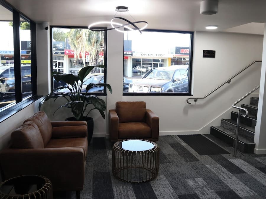 A modern waiting area with a sofa, armchair, and round table, large windows with a view of parked cars, and a staircase.