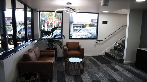 Ein moderner Wartebereich mit Sofa, Sessel und rundem Tisch, große Fenster mit Blick auf parkende Autos und einer Treppe.