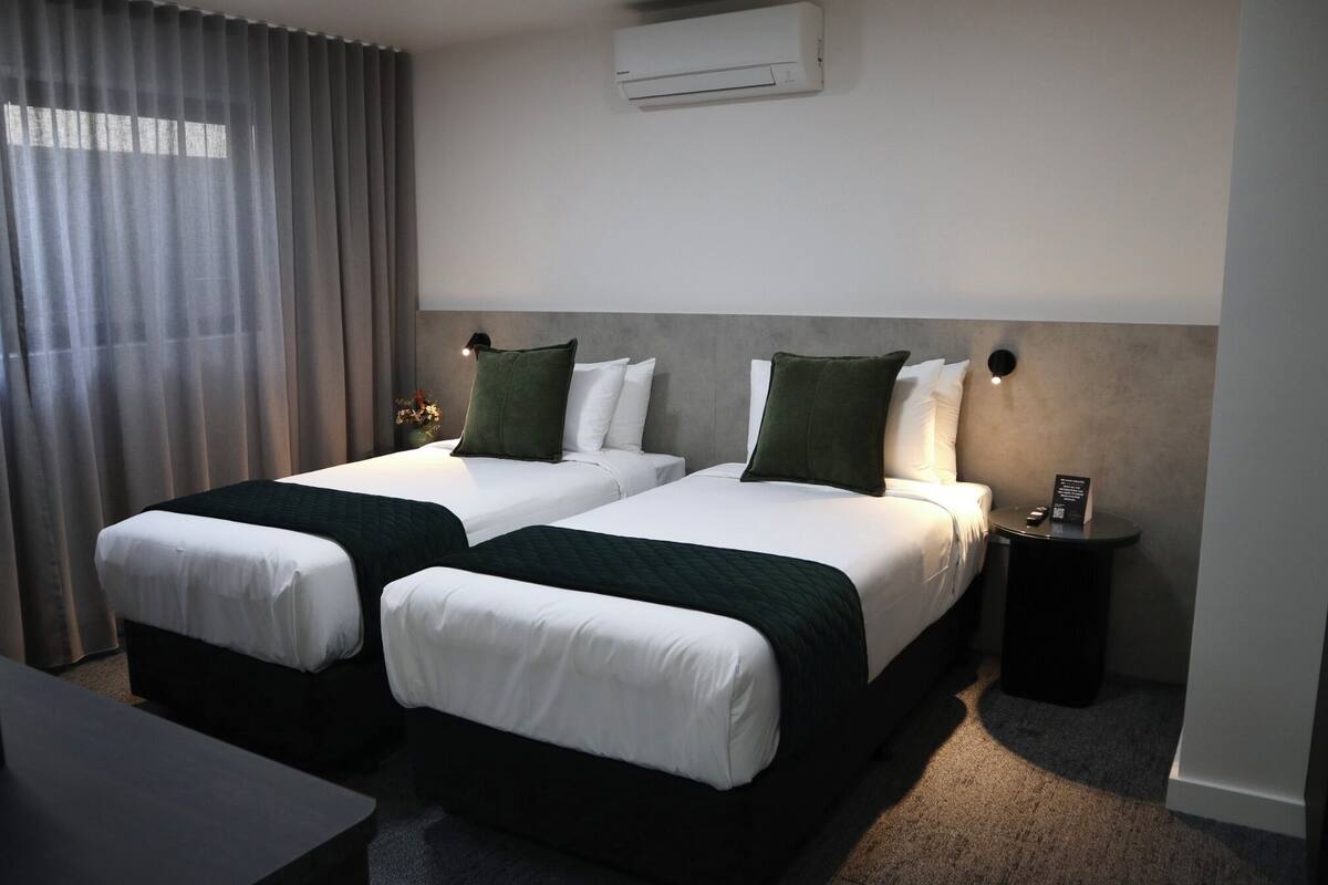 A hotel room with two beds, a desk, a nightstand, an air conditioning unit, and a window with curtains.