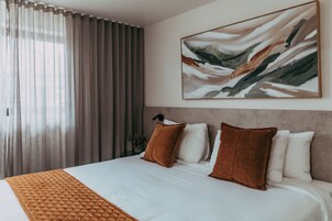 A hotel room with a large bed, a headboard, a painting, and a window with curtains.