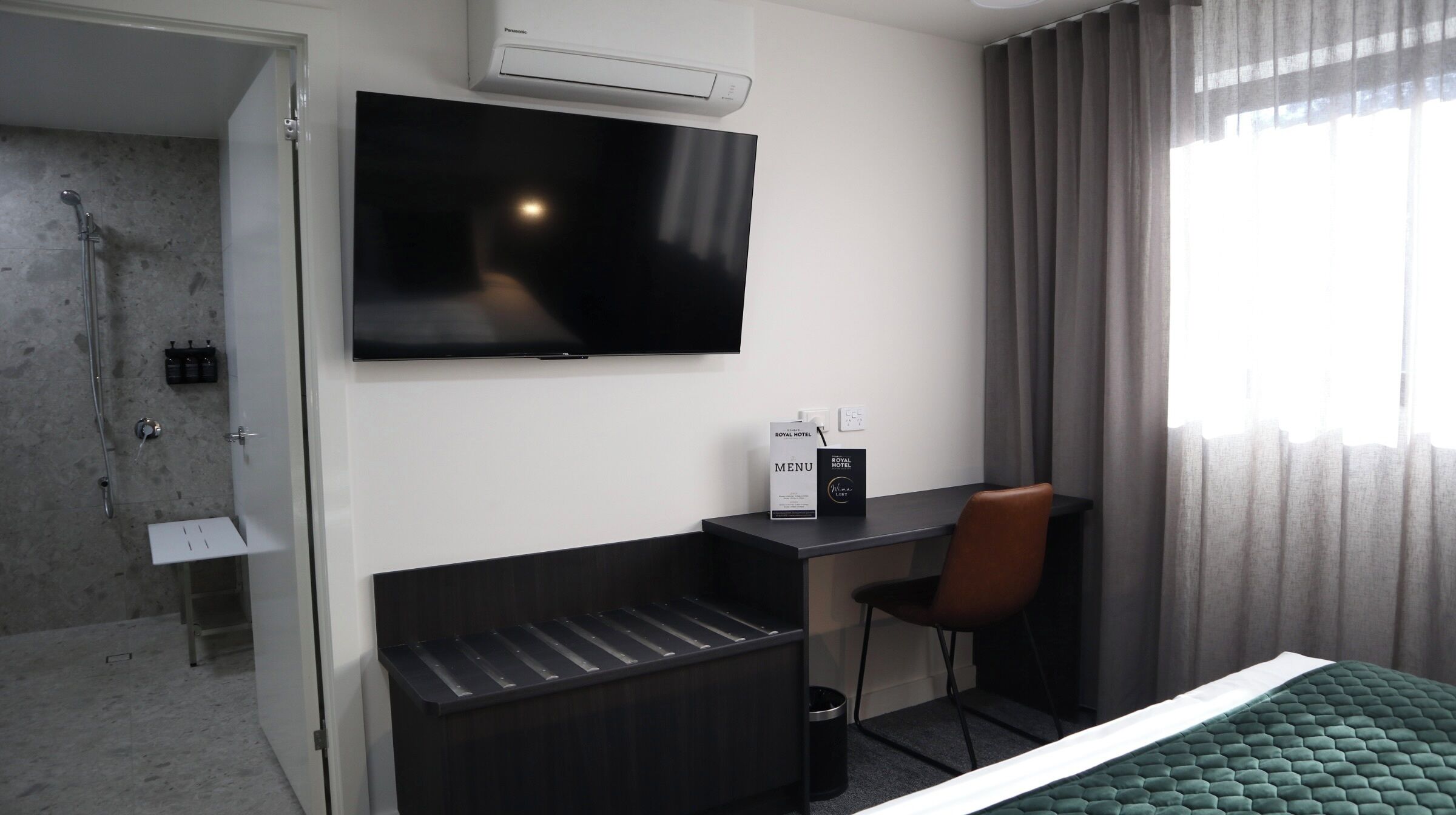 A hotel room with a bed, a desk, a chair, a TV, a shower, and a window with curtains.