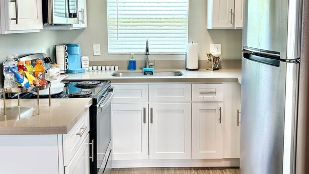 Une cuisine moderne avec des meubles blancs, des appareils en acier inoxydable, une fenêtre avec des stores et un plancher en bois.