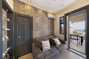 Een moderne woonkamer met een donkere deur, behang met een patroon en een bank met kussens. Er is een glazen plank met decoratieve objecten en een uitzicht op de omgeving door een schuifpui.