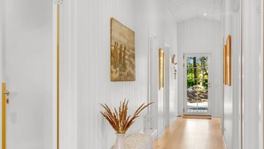 A hallway with white walls, a wooden floor, and a door leading outside. There are framed artworks and a vase with dried plants on a white console table. - "Gedda" -  from the sea by Interhome (Hals)