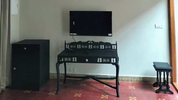 A room with a ceiling fan, a television mounted on the wall, a small table with a decorative backrest, a cabinet, and a small stool. - Svatma Mayavaram (Kuthalam)