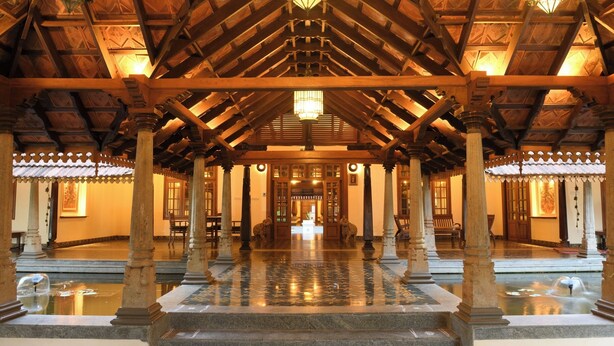 A grand hall with a high wooden ceiling, columns, and a central fountain. - Svatma Mayavaram (Kuthalam)
