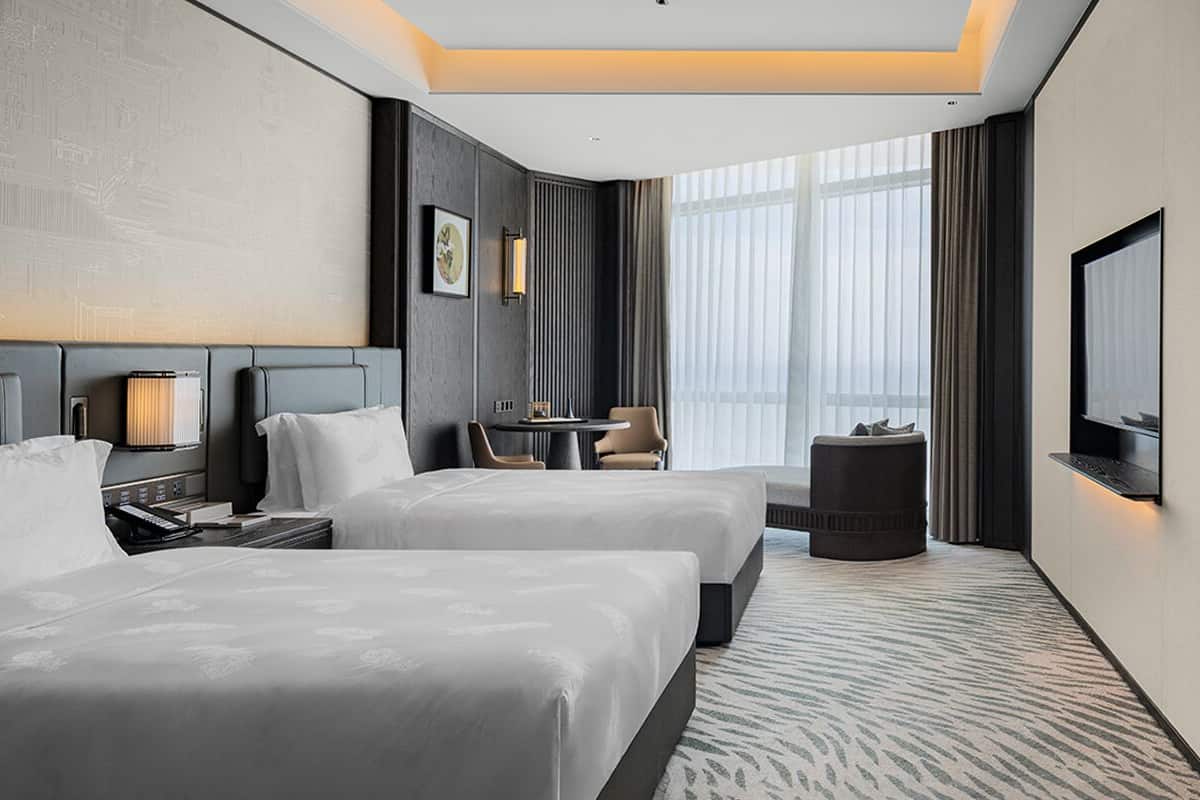 A hotel room with two beds, a desk, a chair, a TV, and a large window with curtains.