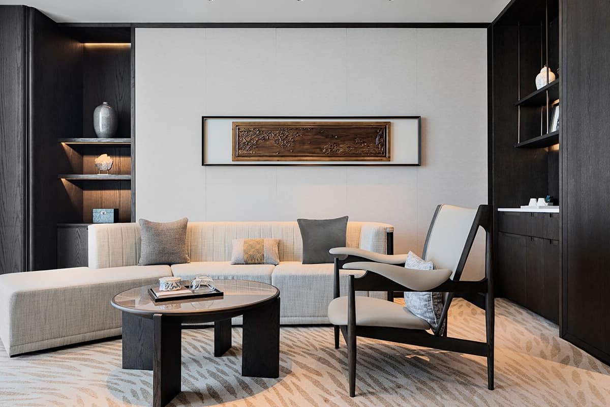 A modern living room with a sectional sofa, a round coffee table, and a minimalist chair. There is a framed artwork on the wall and shelves with decorative items.