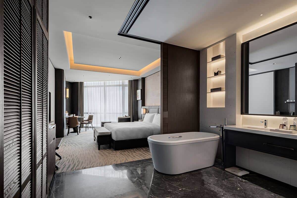 A modern hotel room with a large bed, a bathtub, a vanity with a mirror, and a dining area with a table and chairs.