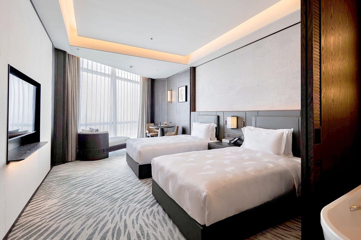 A hotel room with two beds, a large window, a flat-screen TV, and a bathroom with a sink and mirror.