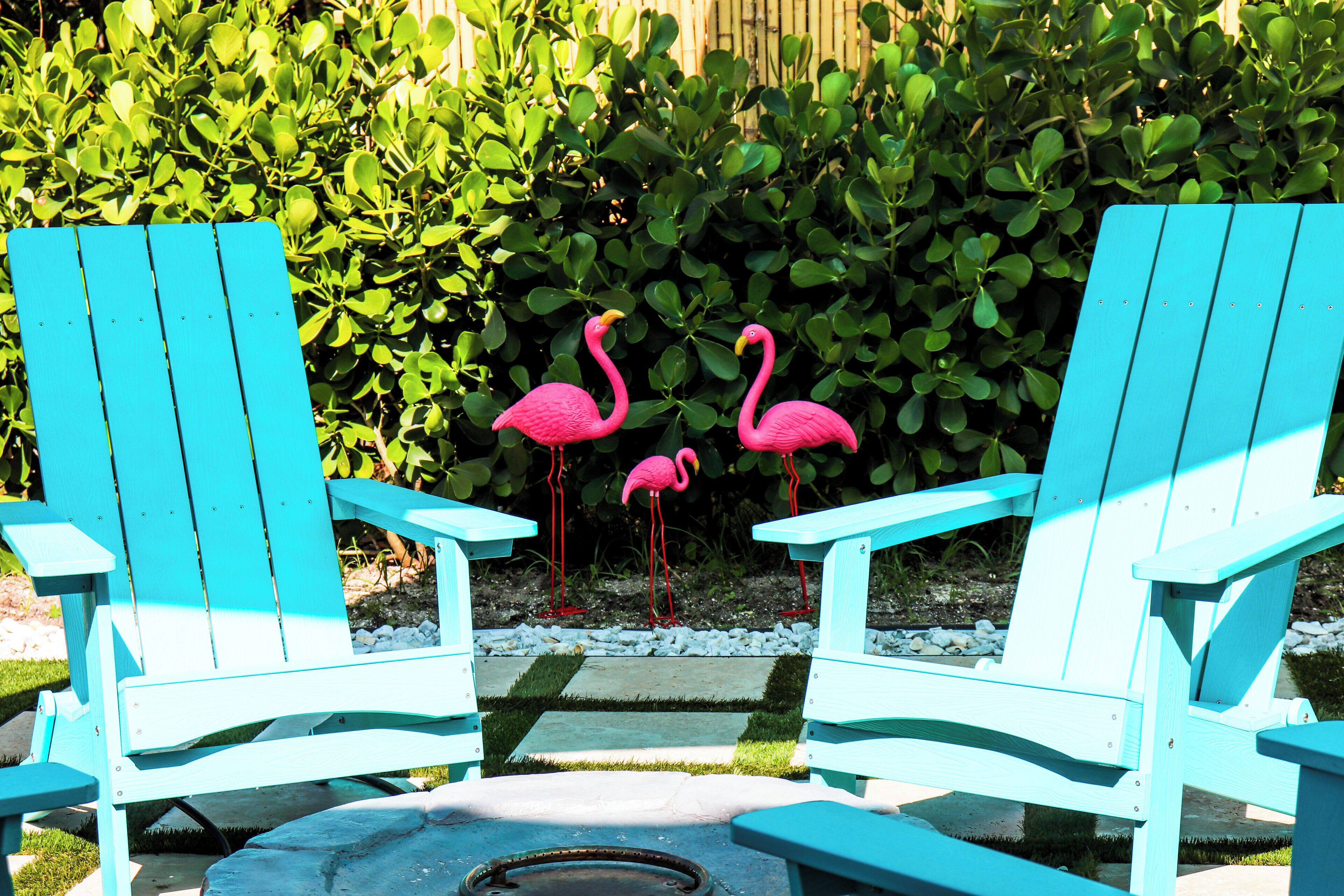 Two turquoise Adirondack chairs facing each other with a small table in between, set against a backdrop of green foliage and a wooden fence.