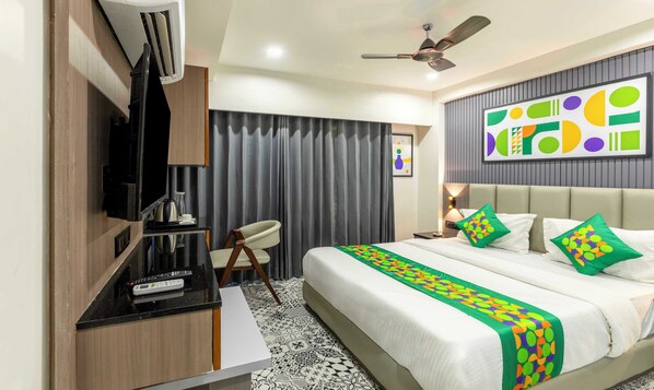 A hotel room with a bed, a desk with a flat-screen TV, a ceiling fan, and a wall with a geometric artwork.