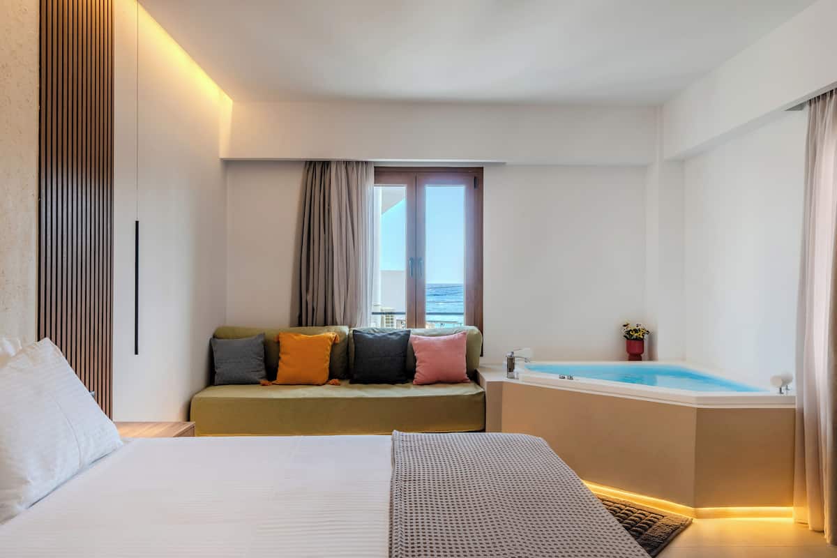 A hotel room with a bed, a sofa with cushions, a window with curtains, and a bathtub.