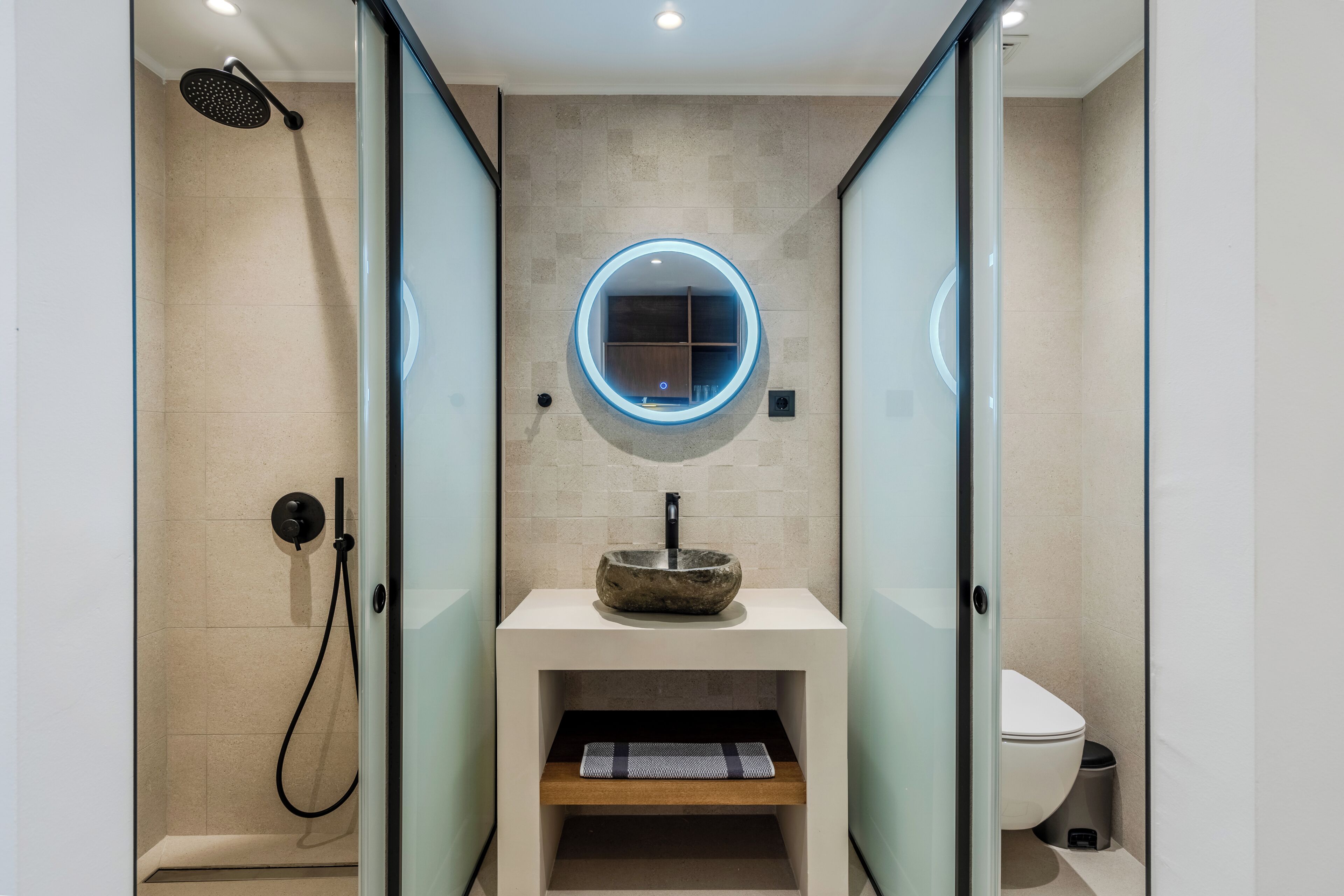 Une salle de bain moderne avec une douche à parois de verre, un miroir rond, un lavabo sur colonne et des toilettes.