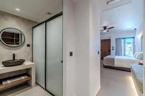 A modern bathroom with a round mirror, a stone basin, and a sliding glass door leading to a bedroom with a bed and a window. - VENTUS Luxury Suites (Sfakia)