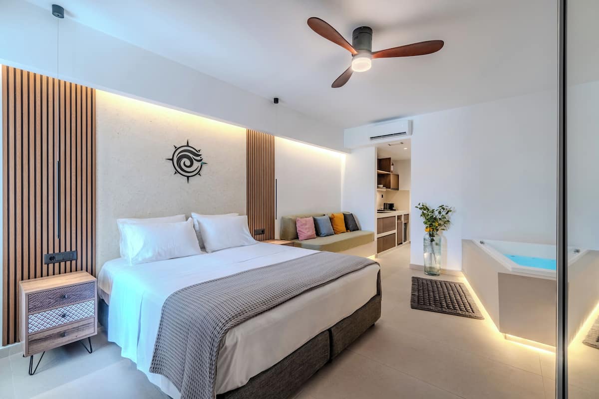 A modern bedroom with a large bed, a ceiling fan, a bedside table, a built-in bench with cushions, a small desk, and a bathtub.