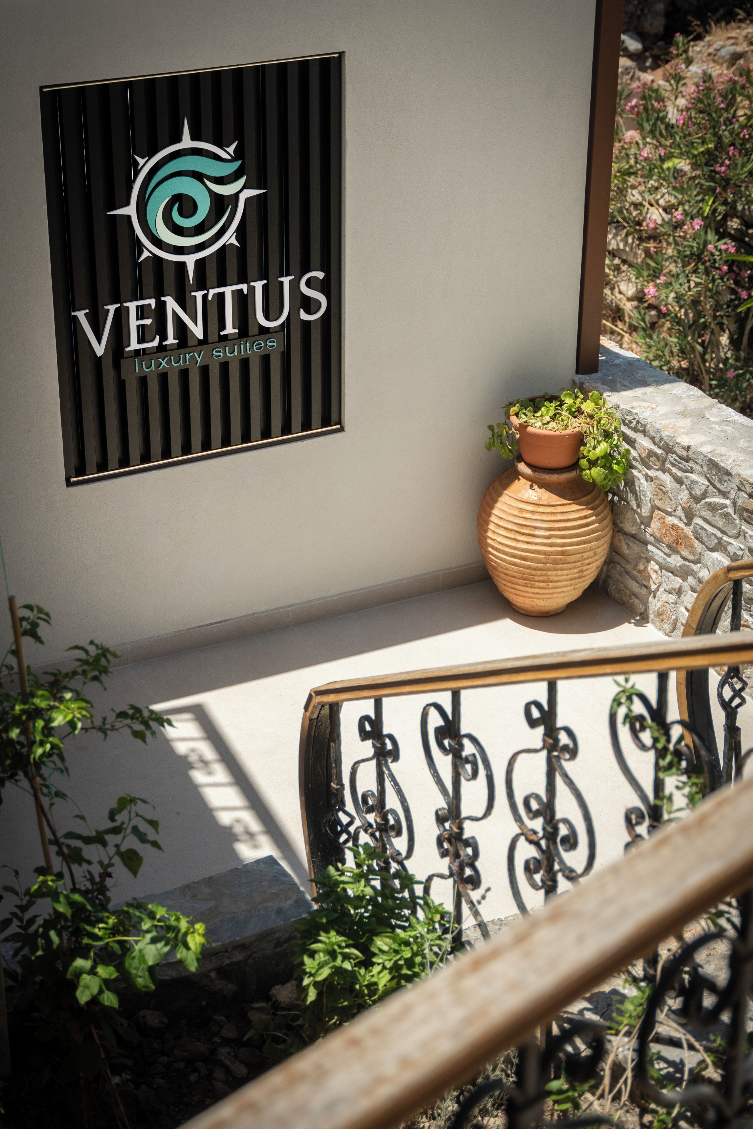A logo for 'VENTUS luxury suites' on a wall, a large terracotta pot with a plant, and a stone wall with climbing plants.