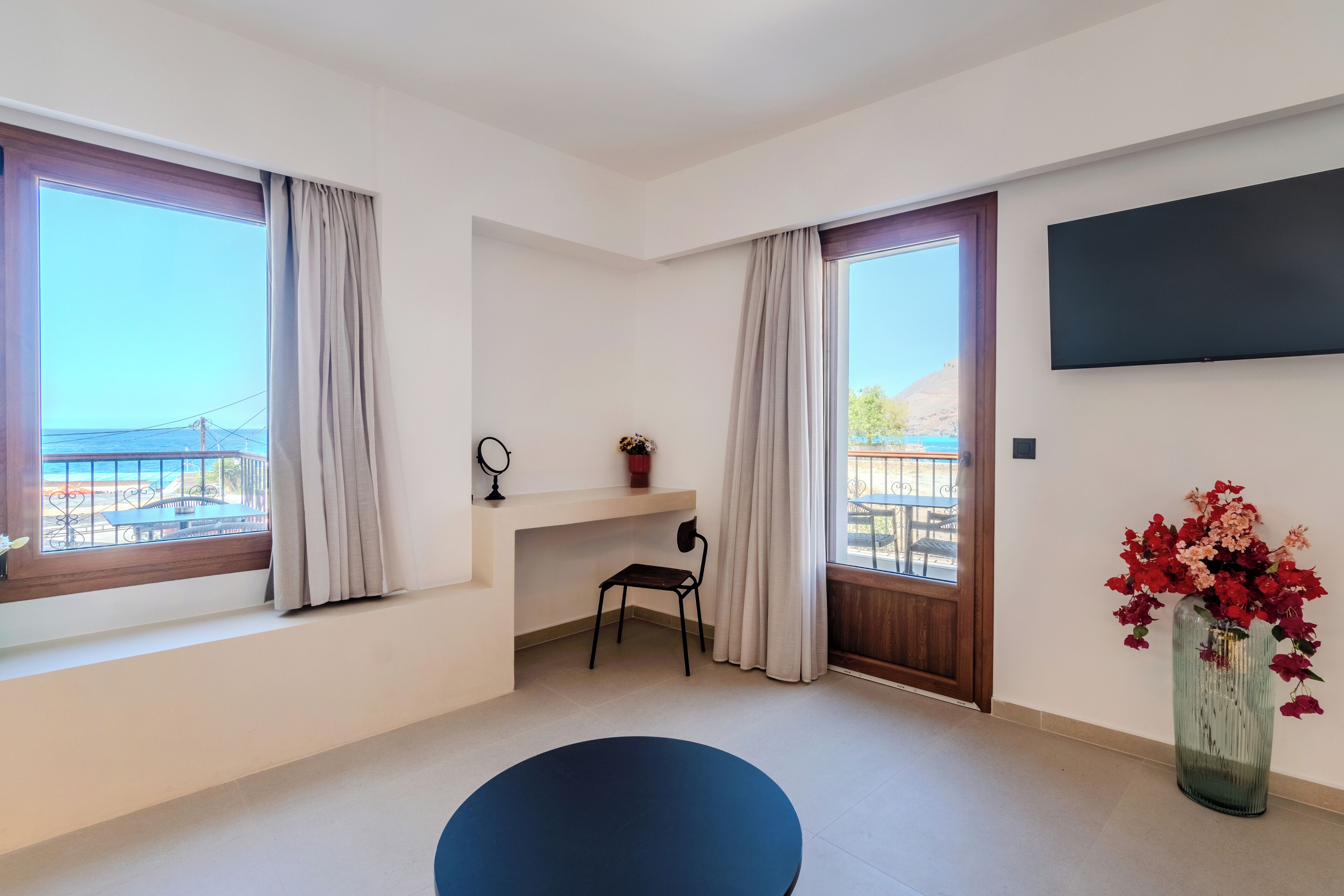 Ein modernes Zimmer mit großem Fenster, einem Wand-Fernseher, einem kleinen Schreibtisch mit Stuhl und einer Vase mit Blumen.