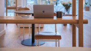 A laptop on a wooden table with a notebook and a mug, a bench, and a view of the outdoors through large windows. - Seaside 9: Modern Studio with Balcony & Ocean View (Cape Town)