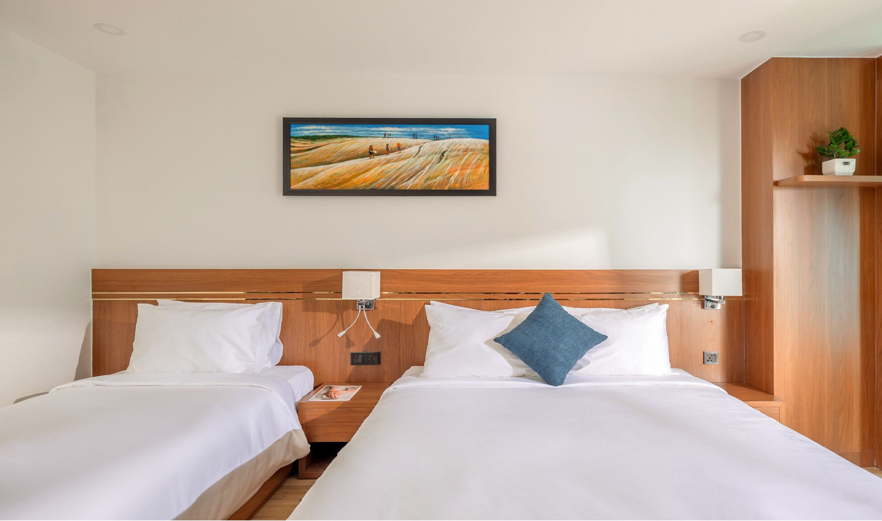 A hotel room with two beds, a wooden headboard, white bedding, a framed painting, bedside lamps, and a small plant on a shelf.