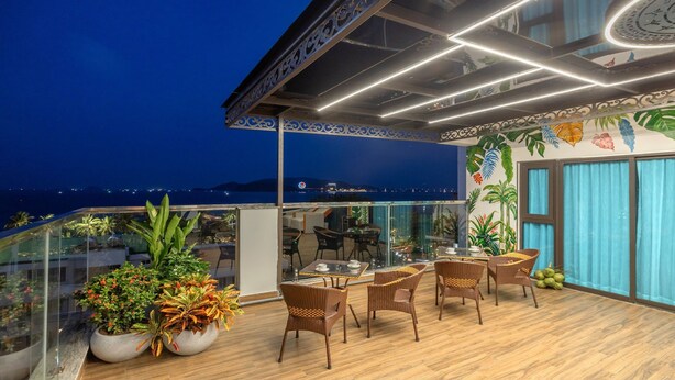 A rooftop terrace with wicker furniture, potted plants, and a view of the sea at night.
