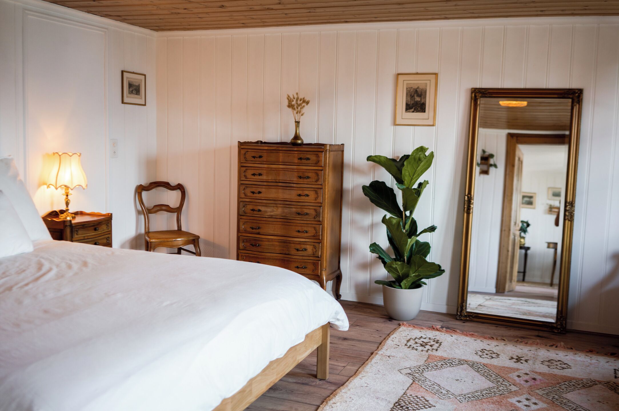 A bedroom with a bed, wooden dresser, chair, potted plant, and a mirror.