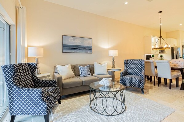 Een woonkamer met een bank, twee gestreepten fauteuils, een salontafel en een eethoek met een kroonluchter.