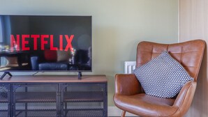 A living room with a television displaying the Netflix logo, a brown leather armchair with a patterned pillow, and a wooden cabinet below the TV. - Two-Bedroom Townhouse (Cardiff)