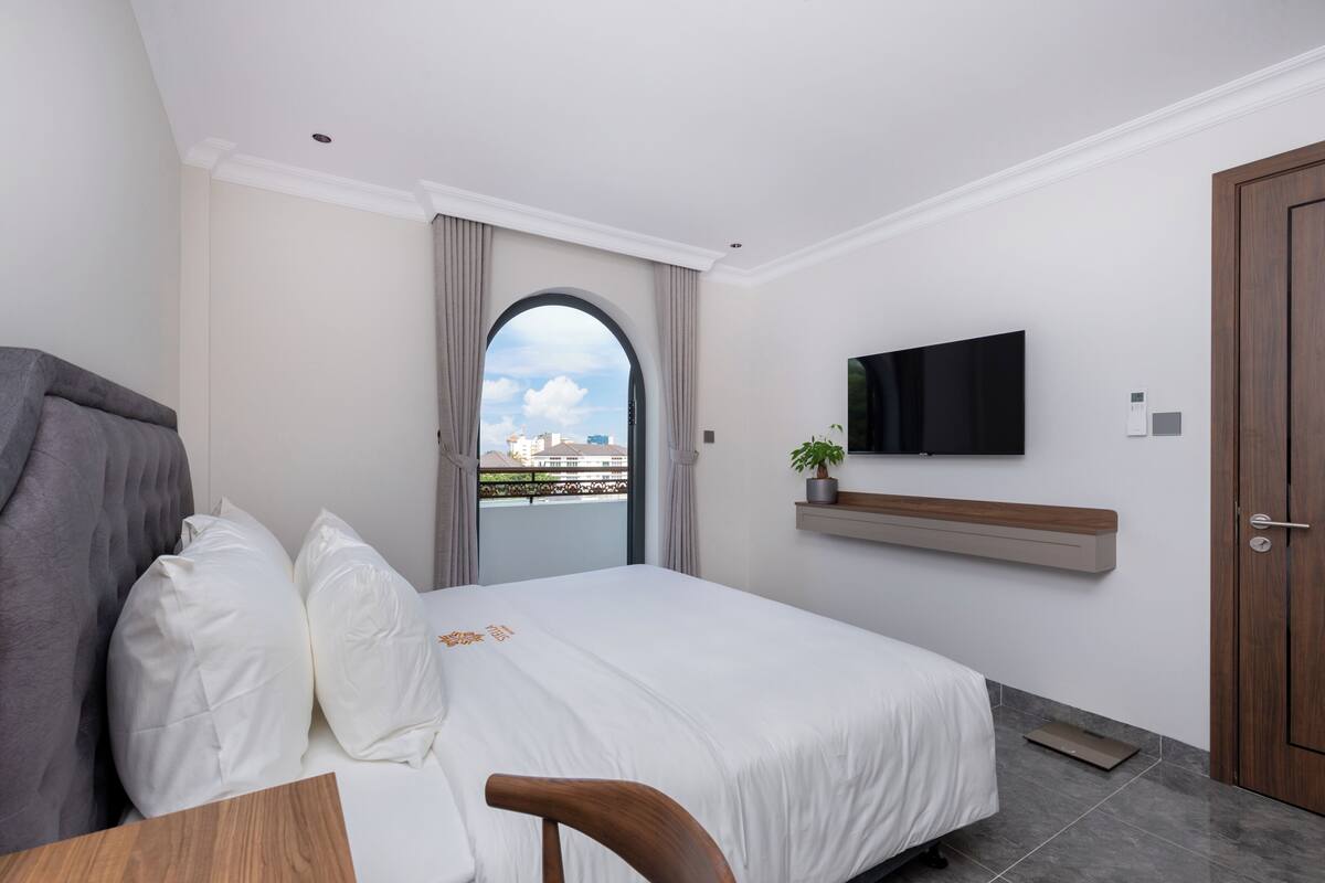 Una habitación de hotel moderna con cama, escritorio, silla, televisión montada en la pared y vista a la ciudad a través de un ventanal arqueado.