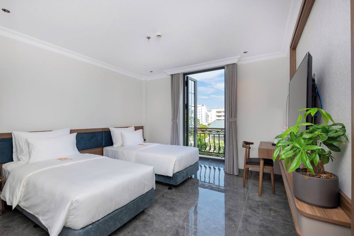 Habitación de hotel con dos camas, un escritorio, una silla, una planta en maceta y un balcón con vistas a edificios.