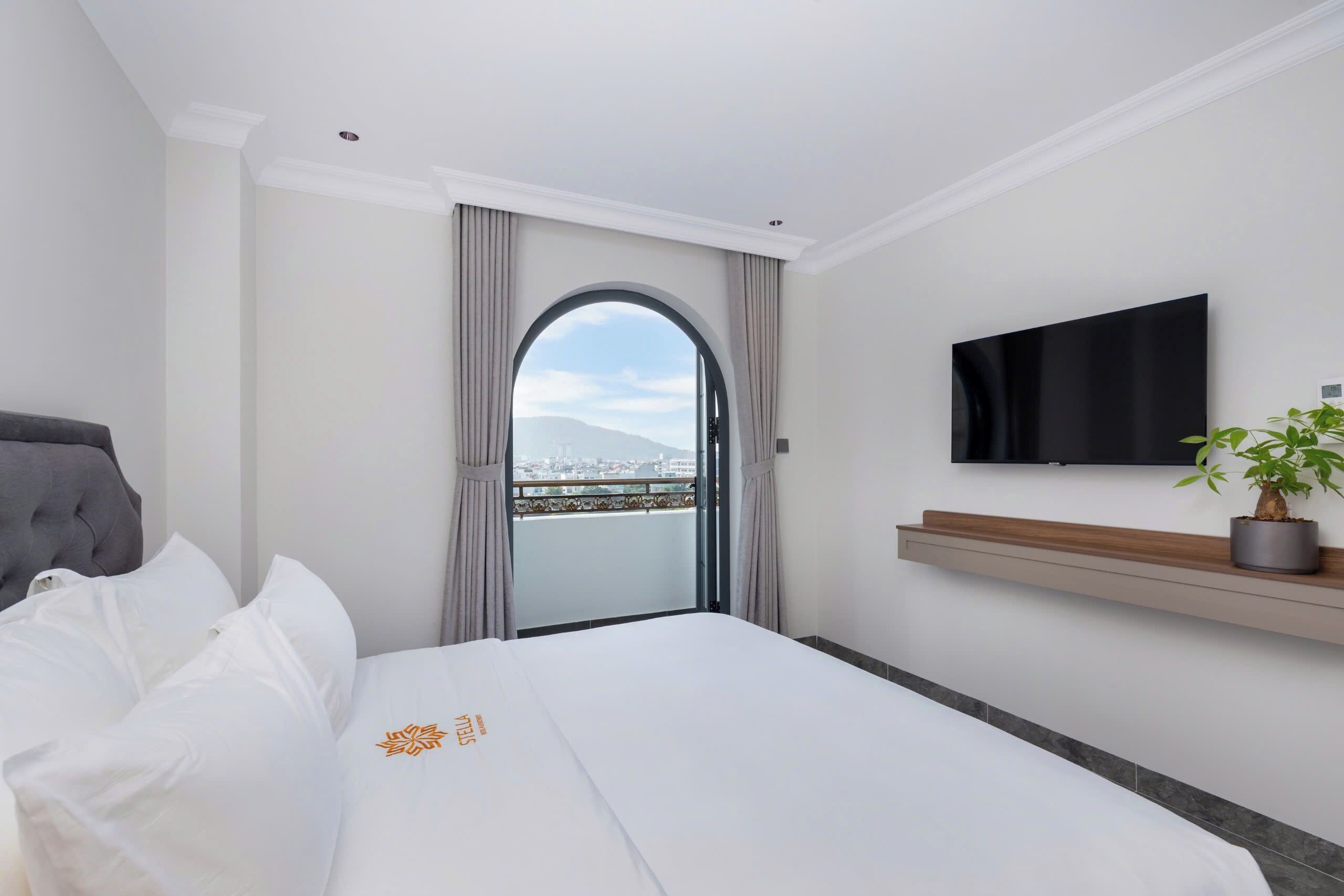 A hotel room with a bed, a TV mounted on the wall, a potted plant, and a view of the city through an arched window.