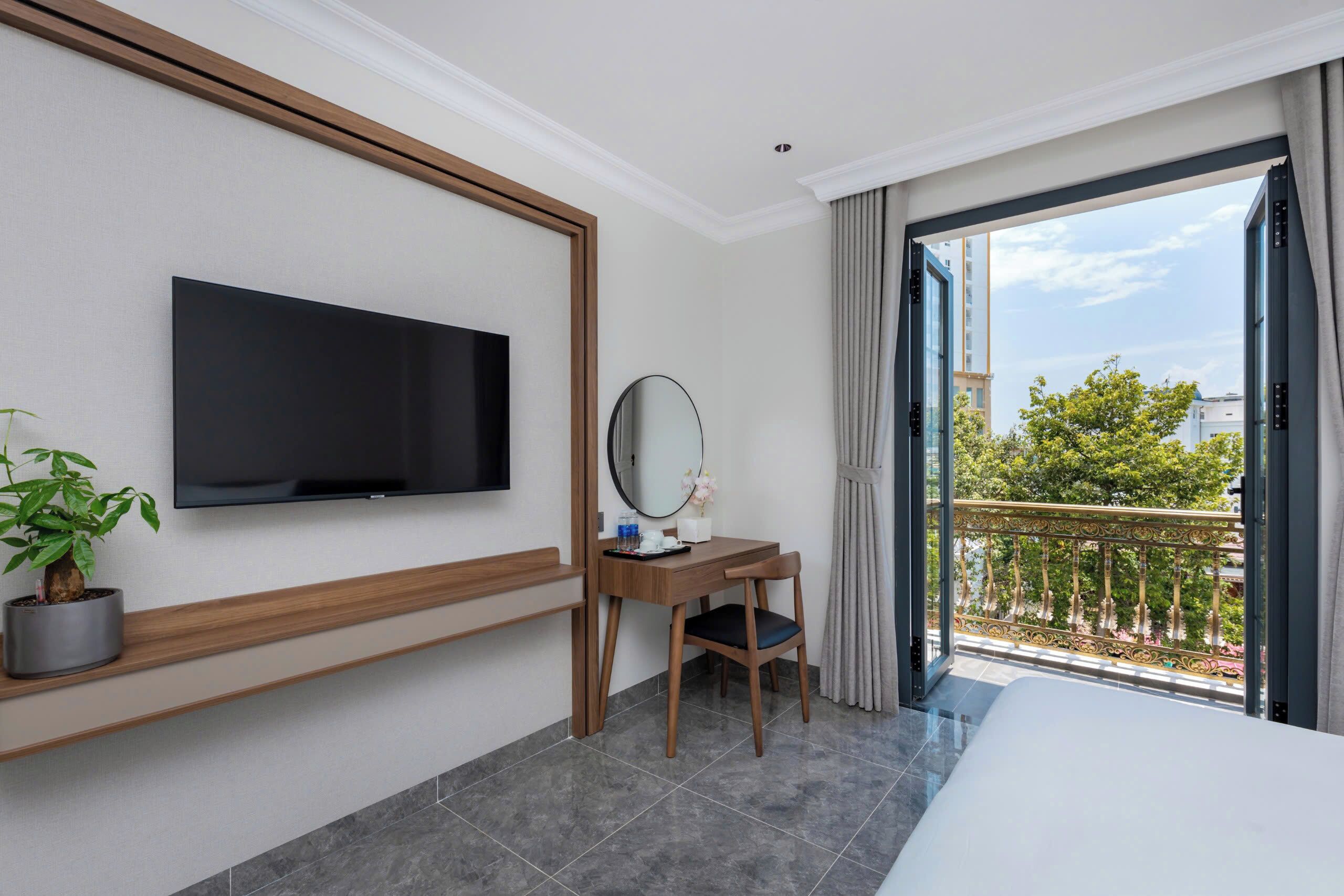 A hotel room with a bed, a TV mounted on the wall, a desk with a chair, a potted plant, a mirror, and a balcony with a view of trees and buildings.