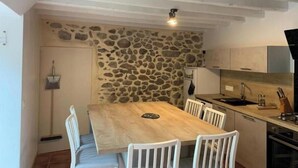 A compact kitchen with a stone wall, wooden table, white chairs, and basic appliances. - Gîte du Cady (Casteil)