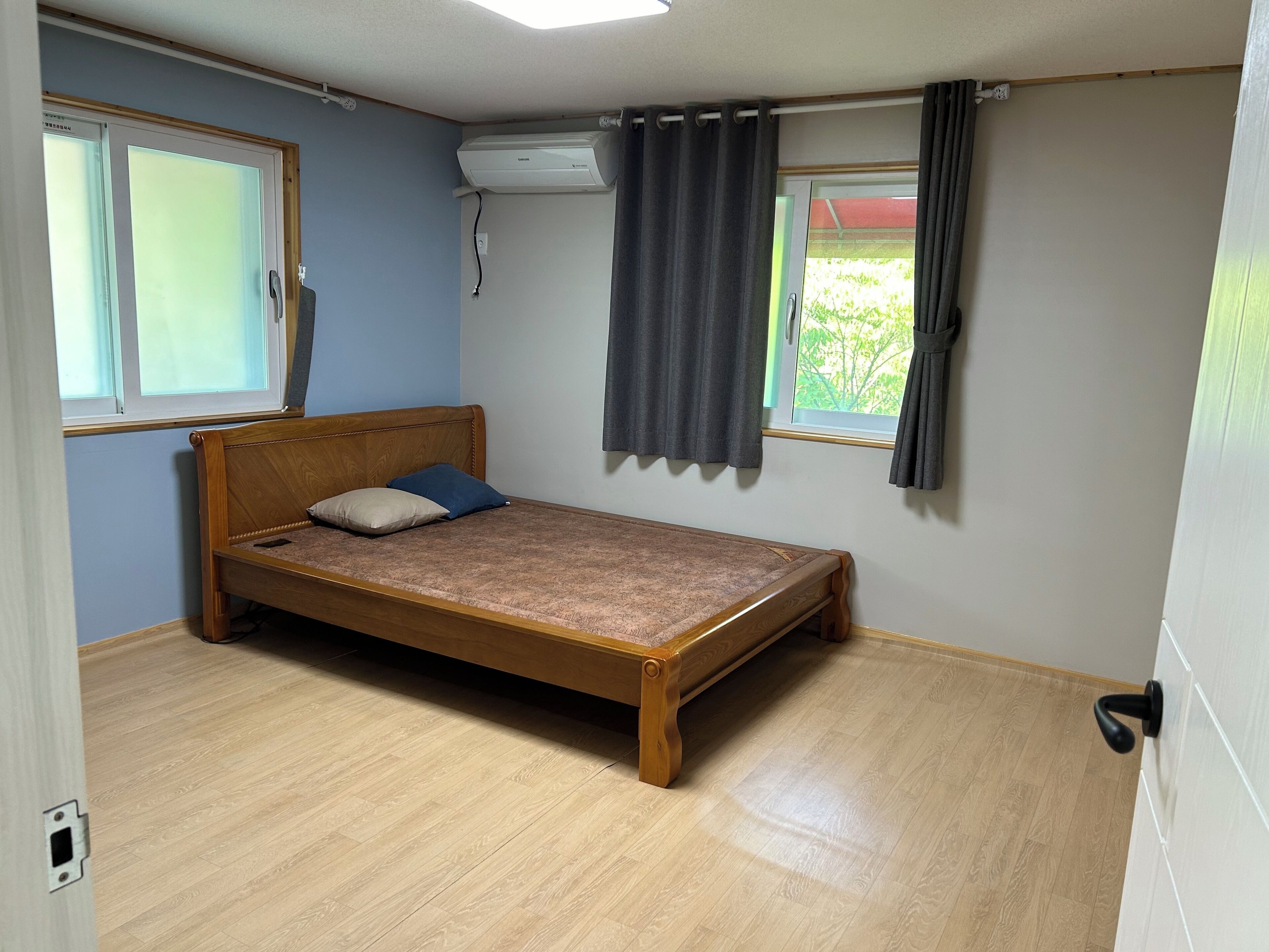 A small, single-bed room with a wooden bed frame, a window with curtains, a ceiling-mounted air conditioning unit, and a light switch on the wall.