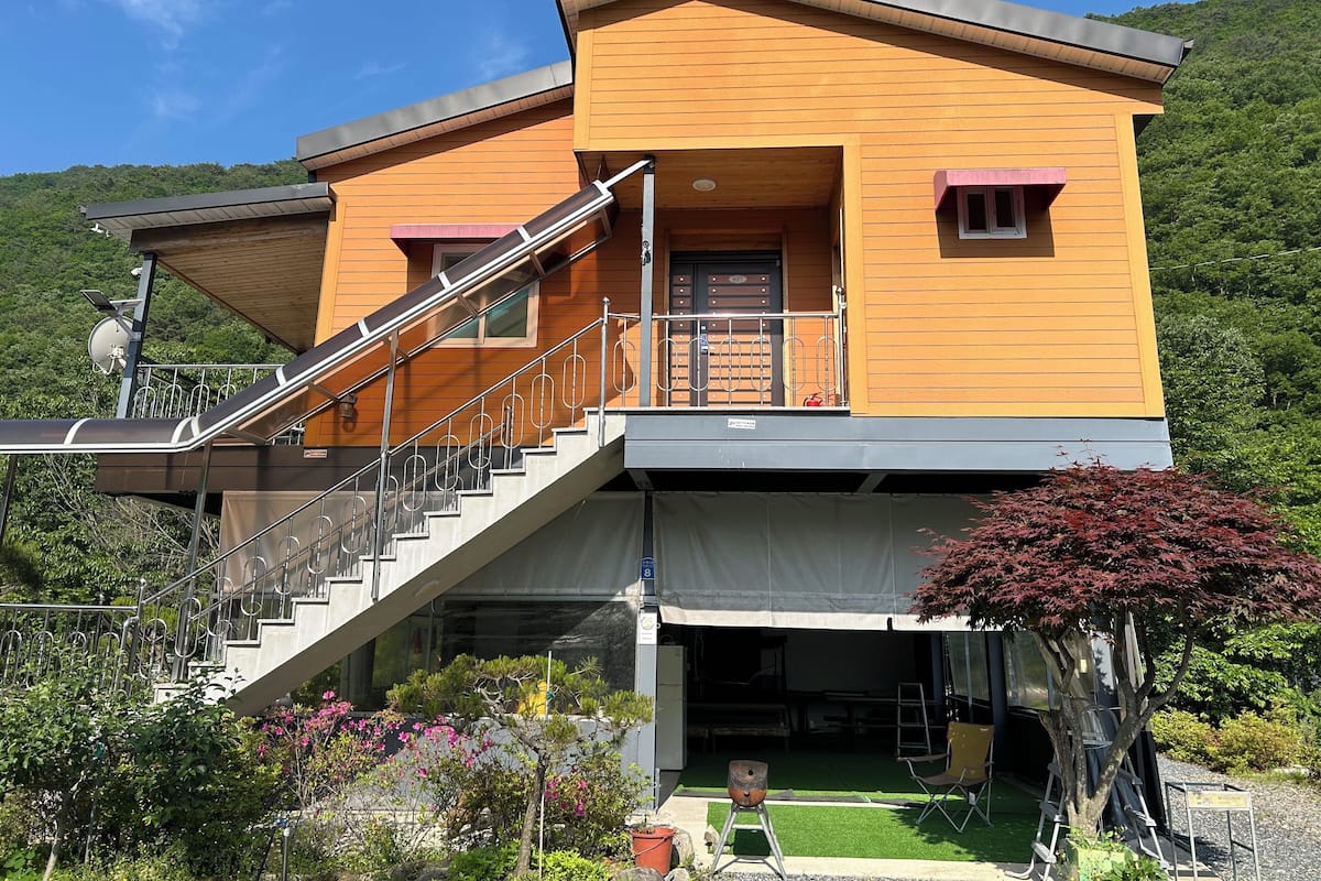 A two-story wooden house with a balcony, a staircase, and a small garden area.