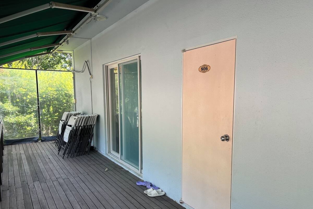 A covered porch with a sliding glass door, a closed door with a number plate, and a row of chairs.