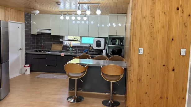 A modern kitchen with wooden walls, a stainless steel refrigerator, a microwave, and a gas stove. There are two bar stools and a small island with a sink. - MAUMAEGOHYANG Pension (Cheongdo)