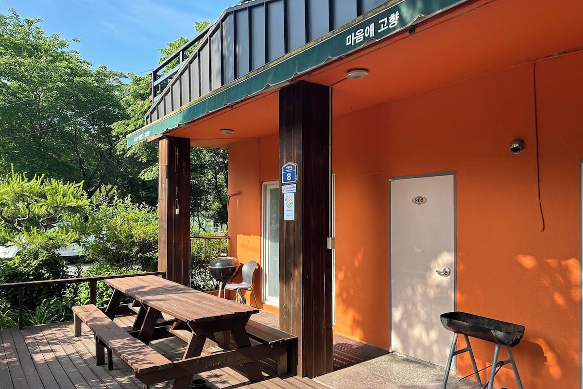 A wooden deck with picnic tables and a barbecue grill, a door labeled "802", and a green awning with Korean text.