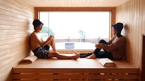Two people sitting on wooden benches in a sauna room with a large window. - AKESUKE (Minamiawaji)