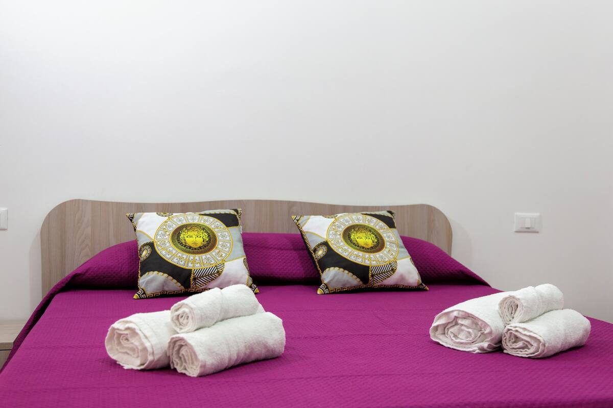 A neatly made bed with purple bedding, two decorative pillows, and rolled towels.