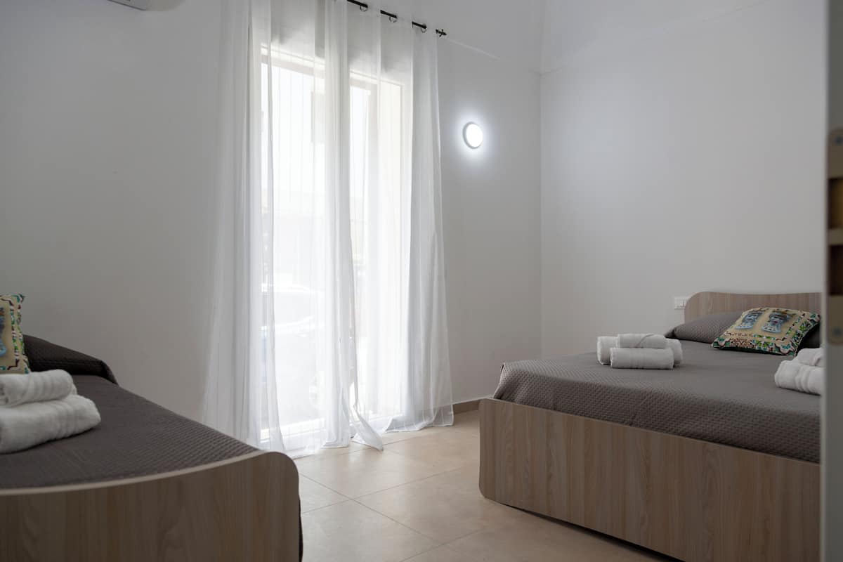 A modern hotel room with two beds, a large window with sheer curtains, and a round light fixture on the wall.