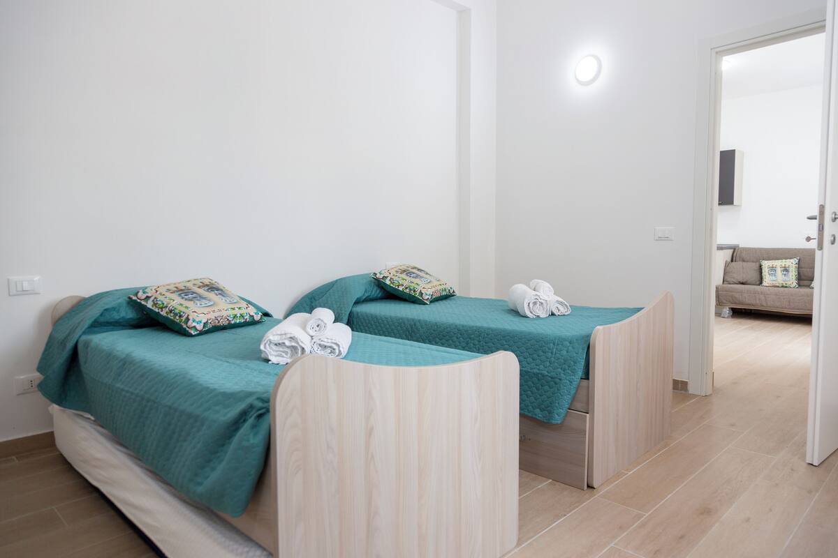 A room with two single beds, each with a teal bedspread and matching pillows, and a wooden headboard. There is a white wall with a light switch and a door leading to another room.