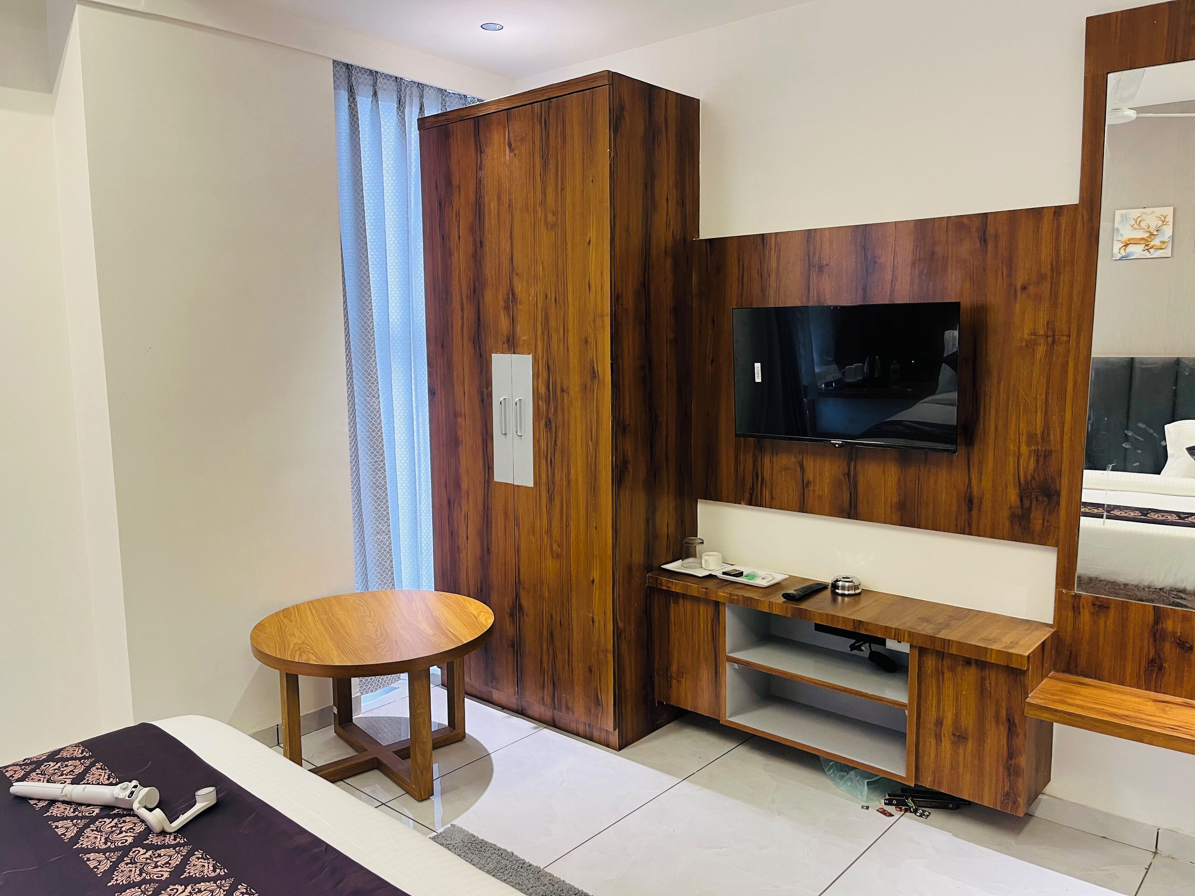 A modern hotel room with a wooden wardrobe, a flat-screen TV, a small round table, and a bed with a patterned bedspread.