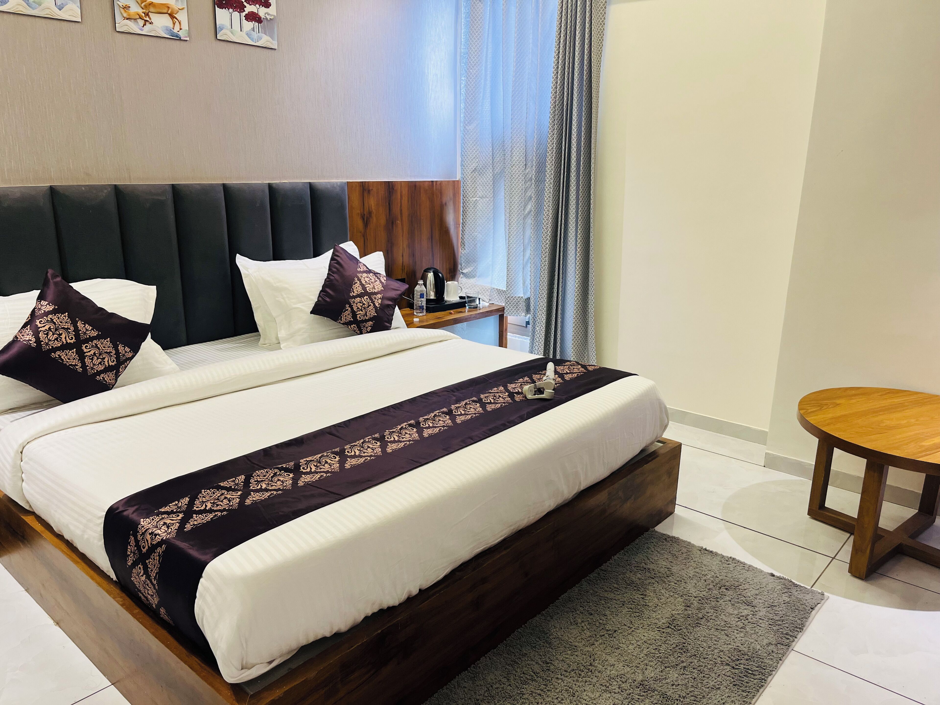A neatly made bed with a dark headboard, white and patterned pillows, and a dark bedspread. There is a small wooden table beside the bed, and a grey rug on the floor.