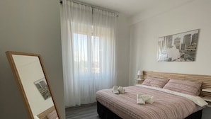 Un dormitorio con cama, una mesita de noche, un espejo, una ventana con cortinas y un cuadro de paisaje urbano en la pared.